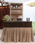 Bar setup with bottles, glasses, and a floral-patterned tablecloth under a tent.